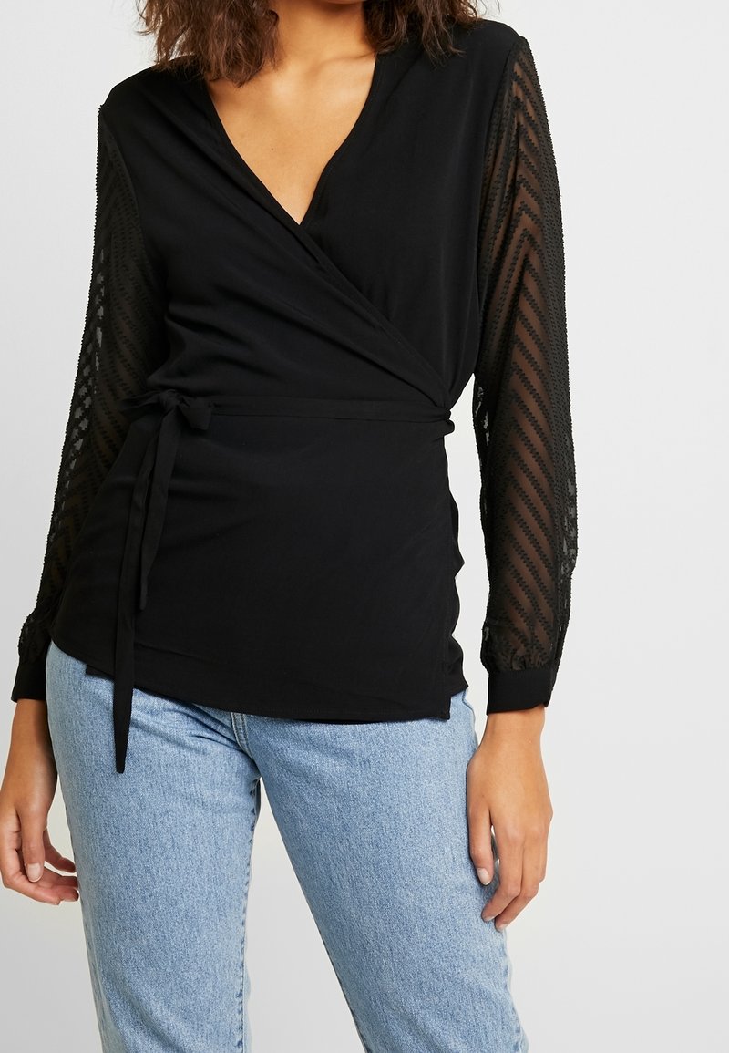 Femme portant un top cache-cœur noir avec des manches longues transparentes et à motifs, et un jean bleu clair, debout devant un fond clair uni.