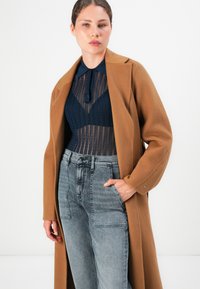 Donna che indossa un cappotto cammello sopra un top in pizzo blu navy con colletto e jeans grigi sbiaditi a vita alta, in piedi con una mano in tasca.