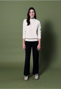 Crème-kleurige, gestructureerde trui met hoge hals, zwart accent, gecombineerd met wijde zwarte broek en lichtbeige schoenen. Groene achtergrond.