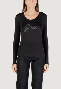 Maglietta nera a maniche lunghe con scollatura a barchetta, con logo "Guess" lucido impreziosito con strass. Abbinata a denim scuro.