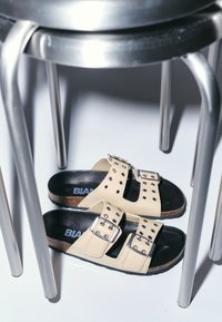 Crème slide sandalen met twee brede banden, zilverkleurige hardware en studaccenten. Kurken voetbed, gecombineerd met een metalen stoelpoten erboven.