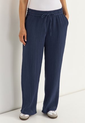 Persoon die marineblauwe, gestructureerde broek met wijde pijpen en trekkoord in de taille draagt, samen met witte sneakers, staand tegen een witte muur.