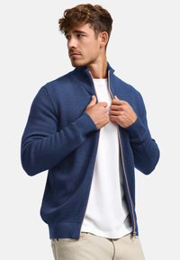 Man draagt een gestructureerde blauwe sweater met rits over een wit T-shirt, past zijn kraag aan, kijkt naar links, staat tegen een effen achtergrond.