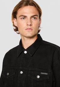 Black denim jacket featuring a pointed collar, front pockets with button closures, and a fabric logo tag on the chest.