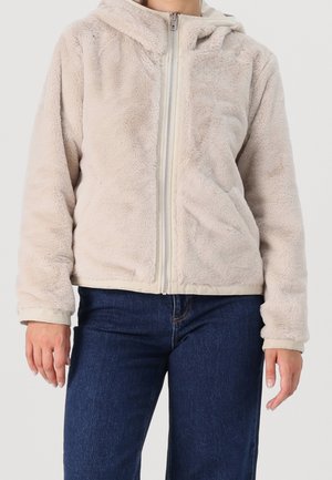 Personne portant une veste polaire beige à fermeture éclair avec capuche et un jean bleu foncé, debout devant un fond uni.