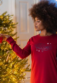 Vrouw in rood shirt met de tekst "Fijne feestdagen" die een rode kerstbal aan een verlichte kerstboom binnenshuis hangt.