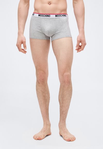 Αρσενικό μοντέλο που φοράει γκρι εσώρουχα Moschino boxer, στέκεται ξυπόλητο πάνω σε λευκό φόντο.