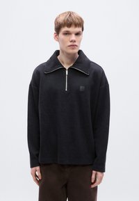 Sweat-shirt noir à col montant avec fermeture éclair. Comprend un petit patch logo et une coupe décontractée. Texture lisse et manches longues.