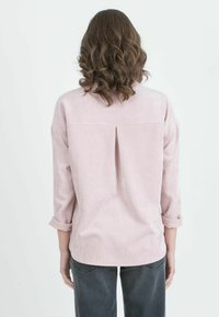 Vaaleanpunainen corduroy-paita kolmella neljänneksellä hihoja, takana pleatti ja kääritty hihansuu. Teksturoitu pinta, jossa on pystysuoria viivoja.