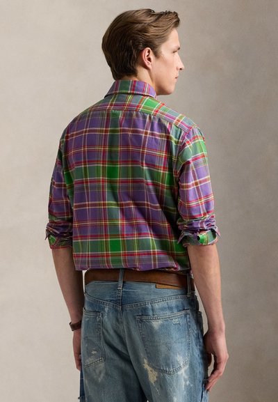Bărbat văzut din profil, purtând o cămașă în carouri verde și violet cu mânecile suflecate și blugi albaștri uzati, pe un fundal simplu.