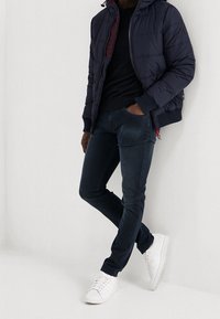 Marinblå puffjacka med huva, svart stickad tröja, mörka jeans och vita sneakers. Jackan har horisontell sömnad och dragkedja.