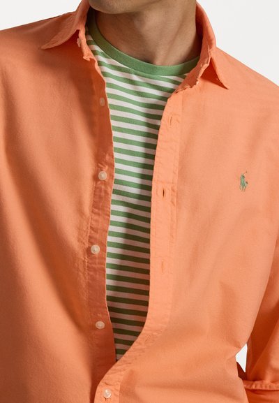 Vaalean oranssi nappipaita, joka on kerroksittain vihreän ja valkoisen raidallisen t-paidan päällä. Paidassa on merkittävä hevosen logoa esittävä brodeeraus.