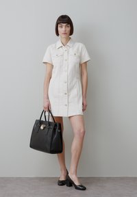 Nainen, jolla on lyhyet tummat hiukset, valkoinen nappilista-mekko, suuri musta käsilaukku ja mustat paksukorkoiset kengät.