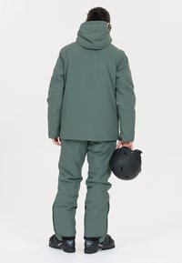 Grön isolerad skidjacka och byxor, med hög krage och åtsittande ärmar. Bäraren håller en svart skidhjälm som visar en elegant och praktisk design.