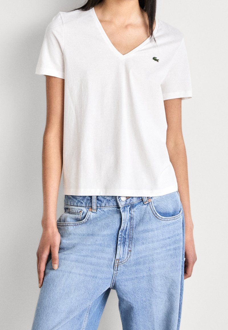 Personne portant un t-shirt blanc uni à col en V avec un petit logo crocodile vert et un jean taille haute bleu clair.