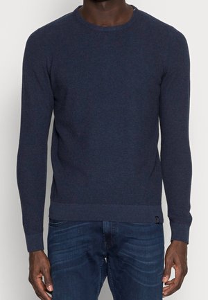 Maglione blu navy con una trama testurizzata, scollatura a girocollo e polsini e orlo a coste. Abbinato a jeans blu scuro, con sottili sfumature e linee.