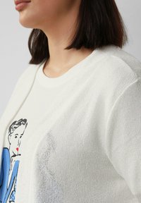 Weißer Strickpullover mit lockerer Passform, der auf der Vorderseite eine Grafik einer Frau in Blau zeigt. Der Pullover betont das strukturierte Material und den runden Ausschnitt.