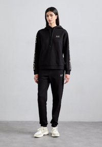 Sweat à capuche noir avec logo, poche kangourou et cordon de serrage. Associé à un jogging noir avec des accents de logo. Les deux sont fabriqués en tissu doux.