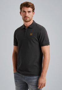 Donkergrijze polo met een ritskraag, korte mouwen en een borstpatch. Heeft een gestructureerde stof en is gecombineerd met lichtblauwe jeans.