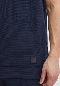 Mørkeblå, tekstureret pullover med en lomme og et kvadratisk mærke. Stoffet har et vævet mønster, der giver en funktionel designdetalje.