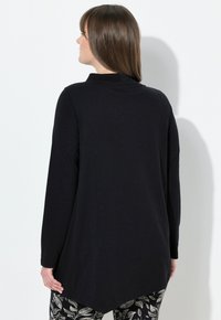 Zwarte lange mouwen top met een hoge kraag, voorzien van een langere achterzoom en een zachte textuur, in combinatie met broeken met een patroon van bladeren.