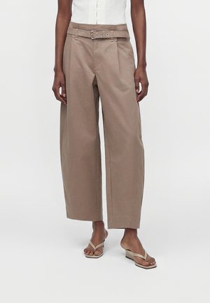 Kvinde iført beige bukser med høj talje og brede ben med bælte, kombineret med beige kilehælssandaler og en hvid ærmeløs top.