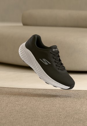 Mustade Skechersi tossud valge tallaga, hõljuvad beežist vaibast kõrgemal beeži diivani ees.