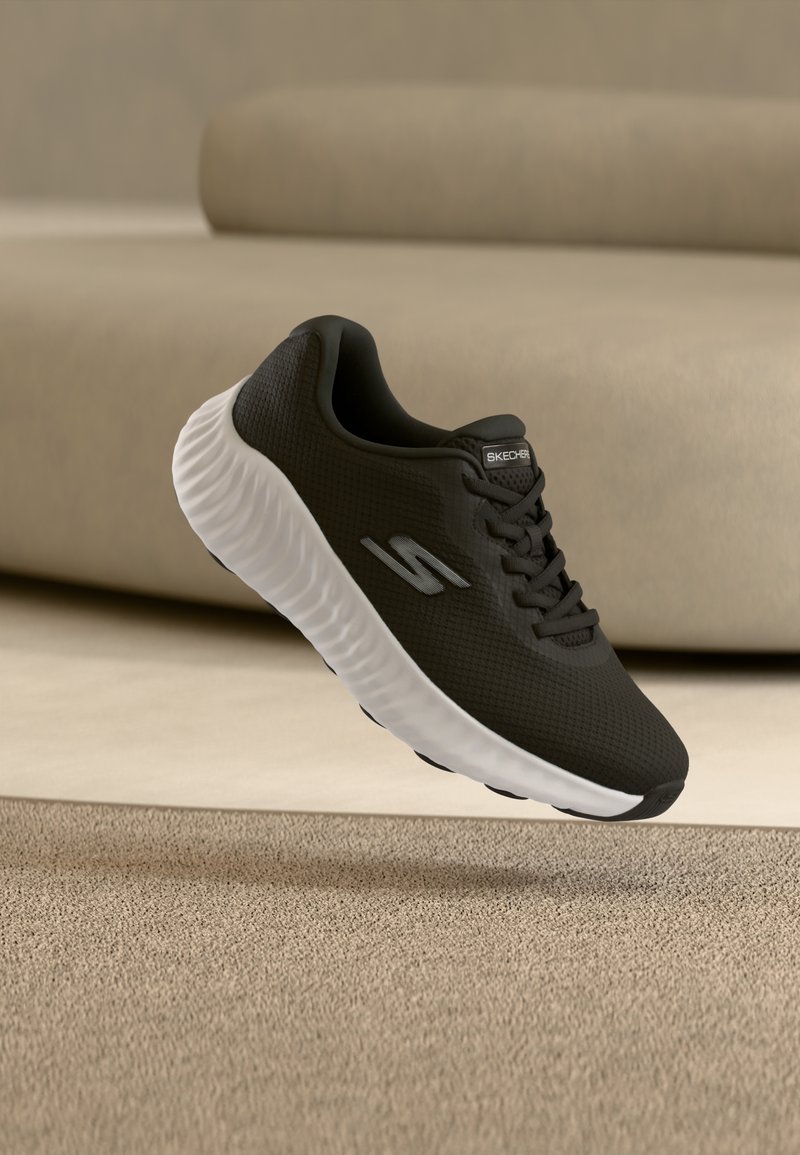 Μαύρο αθλητικό παπούτσι Skechers με λευκή σόλα που αιωρείται πάνω από μπεζ χαλί μπροστά από ένα καφέ καναπέ.