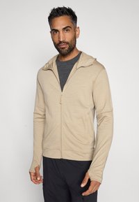 Hanorac crem cu fermoar, cu glugă, mâneci lungi și deschideri pentru degete, realizat dintr-un material moale. Purtați peste o bluză gri, cu buzunare frontale ascunse.