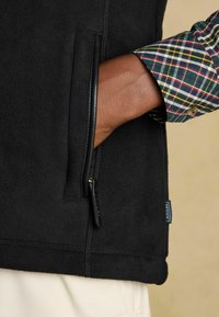 Melnas vilnas veste ar rāvējslēdzēju kabatu. Tam ir fakturēta virsma, gluds rāvējslēdzējs un kontrastējoša svītrainā krekla piedurkne, kas redzama no apakšas.