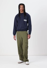 Marineblå hettegenser med hvitt logo, kombinert med løstsittende olivengrønne cargobukser med sidelommer. Hvite joggesko fullfører antrekket.