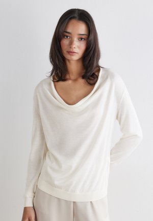 Kvinde med mellemlangt mørkt hår iført en off-white langærmet sweater og lysebeige bukser, stående mod en ensfarvet hvid baggrund.