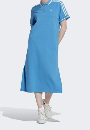 Personne portant une robe polo Adidas bleue avec des rayures blanches sur les manches et le col, associée à des baskets blanches et des chaussettes.