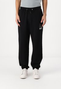 Sorte sweatpants med elastisk talje og manchetter ved anklerne, med en lille grafisk lomme på venstre lår. Båret med hvide sneakers.