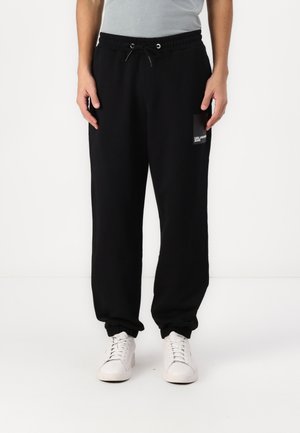 Schwarze Jogginghosen mit einem elastischen Taillenbund und gerippten Knöcheln, ausgestattet mit einer kleinen Grafik-Tasche am linken Oberschenkel. Getragen mit weißen Turnschuhen.