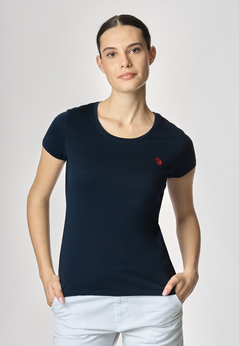 Una donna con capelli lisci indossa una t-shirt aderente blu navy con un piccolo logo rosso, posando con le mani ai fianchi su uno sfondo chiaro.