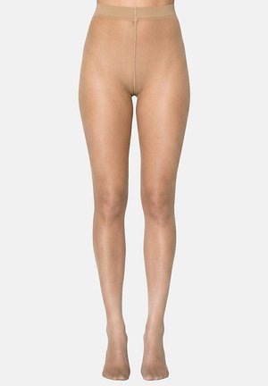 Femme portant des collants nus transparents, debout les pieds rapprochés sur un fond uni.