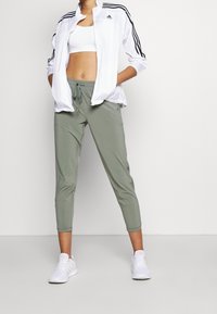 Witte sportjas met ritssluiting en zwarte strepen, gecombineerd met lichtgroene taps toelopende broek en witte sneakers. Gladde stof, comfortabele pasvorm.