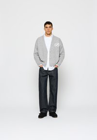 Šedý pletený cardigan s bílým písmenem "B", kombinovaný s bílou košili a tmavými džínami. Outfit je doplněn černými botami.