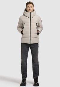 Polstret beige jakke med høj krave og hætte, med sorte lynlåse og to forlommer, parret med mørk denim og sorte sko.