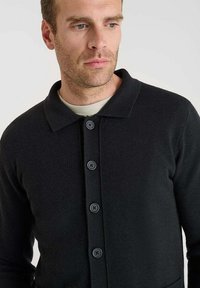 Uomo che indossa un cardigan nero a bottoni sopra una camicia grigia chiara, che guarda leggermente a sinistra su uno sfondo bianco semplice.