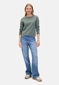 Grüner Baumwollpullover mit langen Ärmeln, kombiniert mit hellblauen ausgestellten Jeans. Die Turnschuhe sind beige mit einer strukturierten Oberfläche.