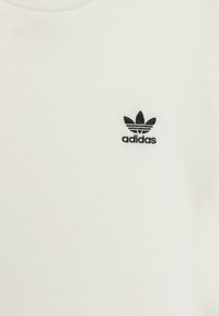 T-shirt en coton blanc présentant un logo Adidas noir avec un design à trois feuilles et des rayures horizontales au-dessus. Texture douce, style col rond.