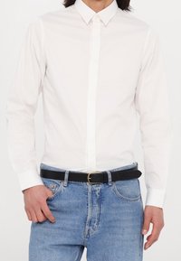 Homme portant une chemise blanche cintrée à manches longues boutonnée rentrée dans un jean bleu avec une ceinture noire et une boucle dorée.