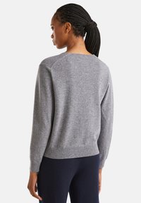 Kobieta z warkoczami spiętymi z tyłu, ubrana w szary sweter z długim rękawem i ciemne spodnie, stoi tyłem na jednolitym białym tle.