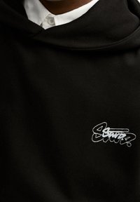 Czarna bluza z kapturem z teksturowanego materiału, z białym haftowanym logo na lewej piersi oraz luźnym kapturem.