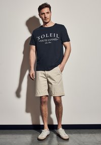 Námořnické tričko s bílým textem, kombinované s béžovými šortkami. Model má na sobě bílé tenisky. Jednoduchý design, měkká textura, volný střih.