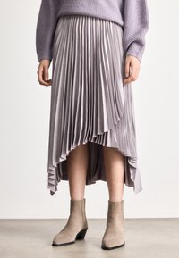 Moteris dėvinti levandų spalvos klostuotą midi sijoną su asimetriniu kraštu, derančią megztinį ir smėlio spalvos aulinukus, stovinti ant pilkos grindų dangos.