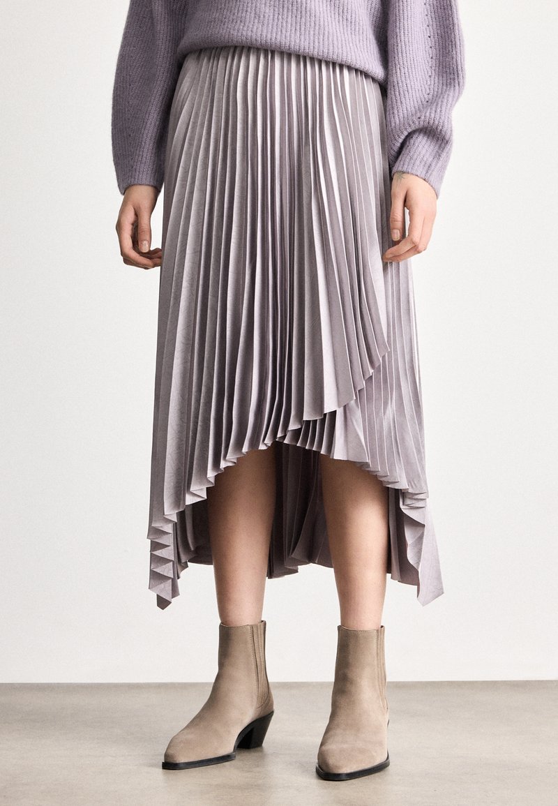 Moteris dėvinti levandų spalvos klostuotą midi sijoną su asimetriniu kraštu, derančią megztinį ir smėlio spalvos aulinukus, stovinti ant pilkos grindų dangos.