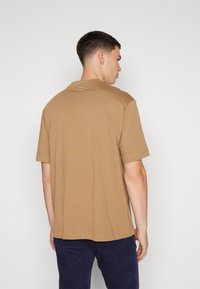 T-shirt in cotone marrone con maniche corte, caratterizzato da un piccolo logo verde sul collo sul retro. Il design è semplice con una vestibilità comoda.